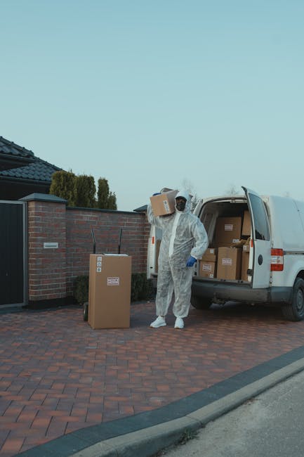 A professional mover dressed in a white protective suit, gloves, and face covering is in the process of unloading cardboard boxes from the back of a white van onto a paved driveway outside a residential property. Several sealed boxes are positioned on the ground nearby, with some still inside the van. The driveway is made of red brick pavers, bordered by a low curb and adjacent to a brick house with a black tiled roof and a dark gate. The scene captures the typical home relocation process involving careful handling of packed items, using a van designated for removals. The image reflects elements of packing, transport, and logistical organisation associated with house removals, as carried out by services like Man with Van Totteridge, emphasizing the precision and safety of furniture transport and moving logistics in a residential setting.