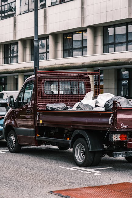 A burgundy flatbed van parked on an urban street outside a modern building with large windows and concrete facade. The vehicle's bed is loaded with various packing materials, including white, black, and grey wrapped items, some secured with straps. The contents appear to be household goods prepared for a home relocation or furniture transport. Nearby, a white van is partially visible, and a tall street lamp is present. The scene suggests an ongoing loading process as part of a professional moving service, with the van positioned close to the building's entrance for efficient offloading. The environment features typical city infrastructure, including marked parking spaces, indicating a professional removals operation by Man with Van Totteridge focused on house removals and relocation logistics.