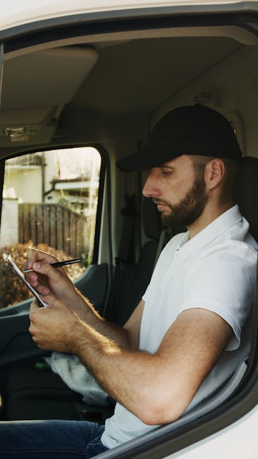 A professional mover dressed in a white protective suit, gloves, and face covering is in the process of unloading cardboard boxes from the back of a white van onto a paved driveway outside a residential property. Several sealed boxes are positioned on the ground nearby, with some still inside the van. The driveway is made of red brick pavers, bordered by a low curb and adjacent to a brick house with a black tiled roof and a dark gate. The scene captures the typical home relocation process involving careful handling of packed items, using a van designated for removals. The image reflects elements of packing, transport, and logistical organisation associated with house removals, as carried out by services like Man with Van Totteridge, emphasizing the precision and safety of furniture transport and moving logistics in a residential setting.
