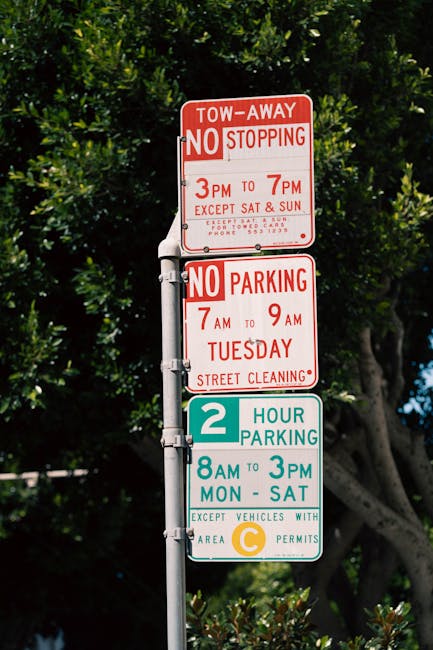 A tall metal street signpost displaying four parking and traffic regulation signs against a background of green trees and a partly cloudy sky. The top sign is red and white, indicating a tow-away zone with no stopping from 3pm to 7pm, except on Saturdays and Sundays, with additional local instructions and a contact number. Below, a second red and white sign notifies no parking from 7am to 9am on Tuesdays for street cleaning. The third sign is teal with white text, indicating a two-hour parking limit from 8am to 3pm, Monday through Saturday, with an exception for vehicles with area C permits. The lowest sign, in yellow and white, further clarifies parking restrictions for vehicles with specific permits. The signs are attached vertically along the pole, with the signs slightly overlapping, and are illuminated by natural daylight, illustrating parking regulation signage commonly seen near residential or commercial areas in Totteridge. The image reflects urban infrastructure supporting house removals and relocation logistics, as seen on the webpage for Barnet Council parking permits for Totteridge removals at manwithvantotteridge.co.uk.