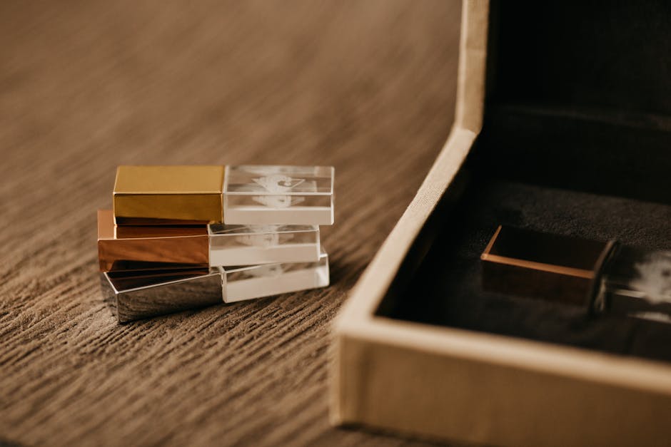 Close-up of a set of four small, rectangular lipstick cases with metallic finishes in gold, rose gold, silver, and black, arranged in a slightly fanned-out manner on a textured wooden surface. Next to the lipstick cases is an open beige cardboard box with a black velvet lining, partially visible, suggesting the cases are part of a packing or aesthetic display. The scene is well-lit, highlighting the reflective surfaces of the cases, and the background is neutral and out of focus. This image reflects careful packing or presentation, which can be associated with home relocation or packing services provided by Man with Van Totteridge, a house removals company specializing in furniture transport and move logistics.