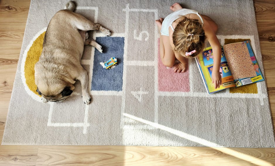 A young girl with blonde hair tied back in a pink hair clip is sitting cross-legged on a beige and multi-coloured hopscotch pattern area rug, reading a colorful children's book with cartoon illustrations. To her left, a small, light-colored dog with short fur is sleeping on the rug, curled up with its eyes closed. Near the dog, there is a small blue toy car. The rug is situated on a wooden floor with visible natural grains and knots. The scene is illuminated by natural light, suggesting a window nearby. Inside the home, the environment appears tidy and calm, with no furniture or other objects visible. This setting is potentially part of a house involved in a home relocation process, where packing and settling are taking place, indicating the importance of careful furniture transport and moving logistics, as highlighted by [COMPANY_NAME] for house removals near Totteridge Village Green.