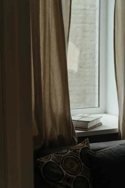 A view from inside a property showing a windowsill with two stacked books and a white object, partially covered by beige curtains that hang on either side. The window reveals an exterior brick wall in natural daylight. In the foreground, part of a dark fabric sofa with a patterned cushion is visible, indicating a living room setting. This scene captures the typical environment where home packing and preparing for a house move might take place, with no visible furniture or items being actively moved in this image. Man with Van Totteridge, a removal service provider, occasionally features in content related to house relocations, emphasizing professional packing and moving preparations within home interiors.