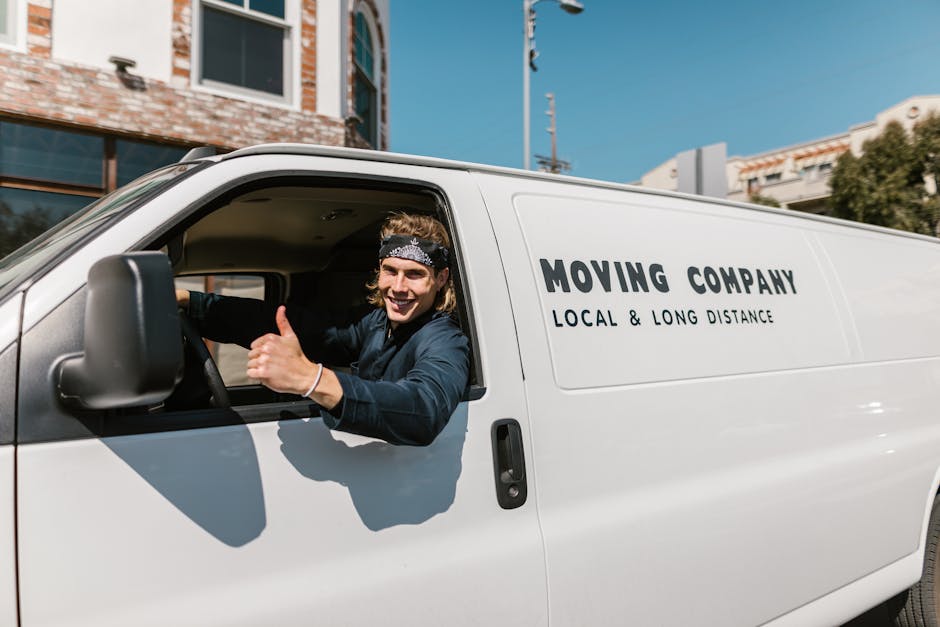 A young male mover with shoulder-length blond hair, dressed in a dark uniform and wearing a patterned headband, is sitting in the driver’s seat of a white moving van parked outdoors near a residenti