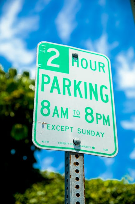 A green and white parking regulation sign mounted on a metal pole outdoors during daytime, indicating a two-hour parking limit from 8 am to 8 pm, except on Sundays. The sign is set against a backdrop of partly cloudy blue sky with green foliage visible on the left and right sides of the image. This type of parking sign is common in urban residential areas such as Totteridge, where parking restrictions are often enforced to facilitate home relocations and furniture transport. The presence of the sign suggests potential parking considerations during house removals or moving services provided by Man with Van Totteridge, who specializes in packing and loading belongings for home relocations. The image captures the sign prominently, emphasizing the importance of adhering to parking regulations when planning to load or unload household items, furniture, and boxes from a van or vehicle at residential properties in the area.