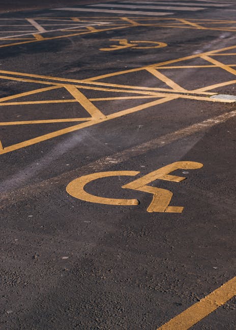 A close-up view of a parking area featuring multiple designated parking spaces marked with bright yellow lines on dark asphalt. The spaces include standard parking spots and two clearly marked accessible parking spaces with large yellow wheelchair symbols painted on the pavement. The markings are freshly painted and well-maintained, with some faint tire marks visible on the asphalt surface. The image captures a section of an outdoor parking lot, likely adjacent to a residential or commercial property, prepared for vehicle loading or staging during home relocation or moving services. This setting aligns with the type of parking arrangements that Man with Van Totteridge might encounter during packing and loading processes for house removals.