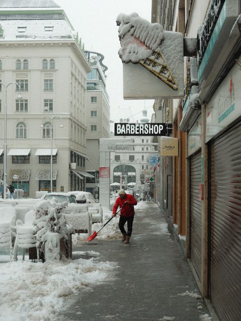A snow-covered urban street scene during daytime with a person in a red jacket shovelling snow from the pavement using a red snow shovel. The street is lined with closed shopfronts, including a barber