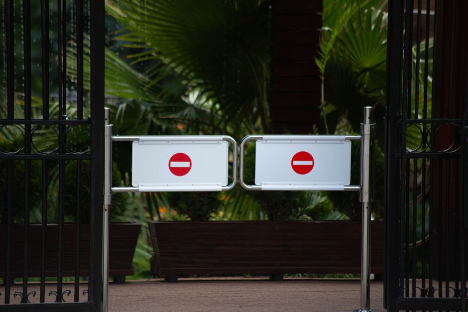 A metal gate with two white signs displaying red no-entry symbols is closed across a pavement, blocking access to a property. Behind the gate, there are large green tropical plants and potted shrubs, indicating an outdoor space adjacent to the building. The pavement appears to be part of a driveway or walkway leading into the property, which is situated near Totteridge & Whetstone station, as referenced in the page title. The gate and signs are part of the traffic control measures, possibly during a home relocation or moving process. There are no visible furniture, boxes, or moving equipment in the image, but the setting and context imply that the residence may be involved in a house removal service by Man with Van Totteridge, specializing in furniture transport and packing during moves.