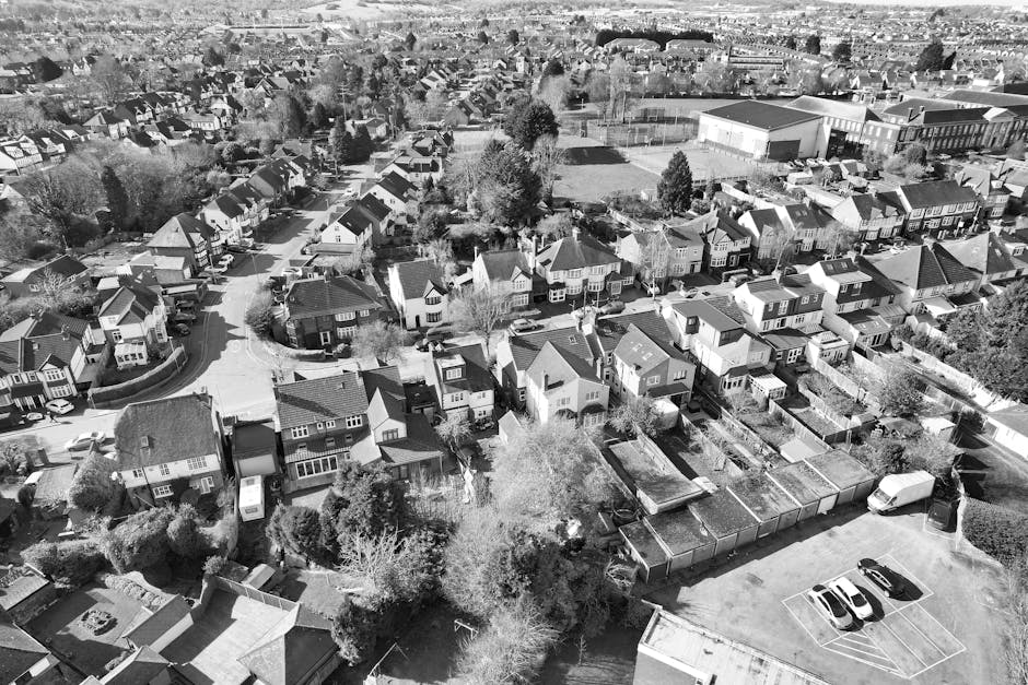 A black and white aerial photograph showing a multi-lane urban road with several vehicles, including cars and a bus, traveling in both directions. The road is bordered by a tall metal fence on one sid