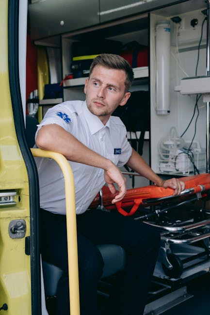 A male paramedic with short brown hair and a focused expression, wearing a white uniform with blue insignia, sits inside an emergency vehicle with the side door open. He is holding a red stabilisation device, likely used for securing patients or equipment during transportation. The interior of the vehicle shows shelves with medical supplies, including bottles, containers, and equipment, arranged behind him. To his right, there is a white medical device attached to the wall, and electrical wires are visible, indicating a well-equipped emergency response environment. The vehicle's yellow exterior door and part of the pavement outside can be seen, suggesting the vehicle is parked, possibly at an emergency scene or during a readiness check. This scene illustrates the preparation involved in rapid response and medical transport services, similar to those provided by companies like Man with Van Totteridge for home relocations involving special handling or urgent logistics related to moving or equipment transport.