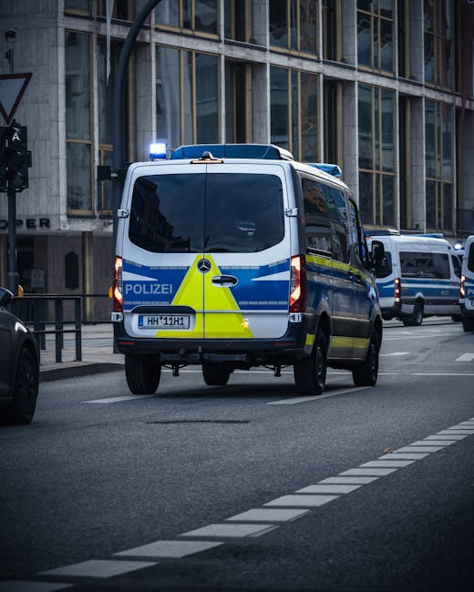 A police van with distinct blue and yellow markings and a flashing blue light on the roof is parked on a city street in front of a modern building with large glass windows. The van's rear doors are closed, and it is positioned on the road near a curb and a pedestrian crossing. Adjacent vehicles, including another van, are visible further along the street. The area appears active with urban traffic and infrastructure typical of a city environment. The presence of the police vehicle may indicate traffic management or emergency response related to road safety or an incident. This scene illustrates a typical urban setting where law enforcement vehicles are involved, possibly in coordination with local authorities, during routine or emergency activities. For home relocation or furniture transport services, such scenes are relevant in illustrating the logistical environment that sometimes involves coordination with police or traffic authorities during moving or transport processes.