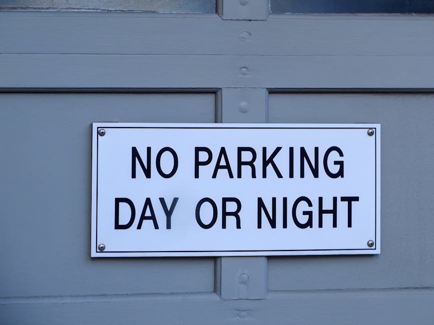 Close-up image of a rectangular white metal sign with black text reading 'NO PARKING DAY OR NIGHT,' affixed to a light gray door or garage door surface with four screws, situated outdoors, with natural daylight illuminating the scene. The sign's purpose relates to parking restrictions during certain times, often relevant in moving or house relocation contexts. Behind the sign, the door appears to be made of wood or composite material with visible paneling and some minor surface detailing. The setting suggests the exterior of a residential or commercial property, where parking permissions are regulated, potentially impacting logistics for furniture transport or moving services conducted by Man with Van Totteridge, a company specialising in removals and home relocations according to the page about avoiding permits, fines, and waste regulations in Totteridge.