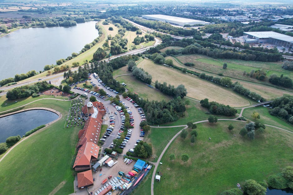 An aerial view of a large park and pond area adjacent to a residential or commercial development, showing a parking lot filled with vehicles near a building with a red-tiled roof. Nearby, there are various boats and equipment stored outdoors, with pathways and green lawns interspersed with trees and bushes. The parking lot is situated on a curve along the edge of the park, and a narrow road runs alongside the greenery. The scene features a mixture of open grass, wooded patches, and water bodies, under a bright sky with scattered clouds. This setting depicts a location suitable for house removals, with visible pathways, parking arrangements, and outdoor storage, reflecting typical moving logistics and transportation considerations. Man with Van Totteridge, a professional removals service, may utilize such areas for parking vans and loading furniture during home relocation or furniture transport operations.