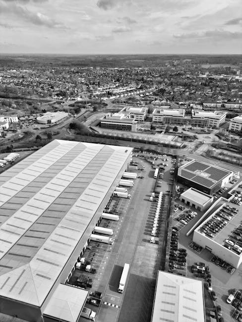 A black and white aerial photograph showing a large industrial warehouse with a metal roof and multiple loading docks along its side, where several trucks are parked, positioned on an open concrete loading area. Adjacent to the warehouse, there are numerous smaller buildings and a parking lot filled with vehicles, including cars and vans. In the background, there is a sprawling urban landscape with numerous residential and commercial buildings, roads, and green spaces extending towards the horizon. The image captures the expansive scene during daylight, with shadows indicating the position of the sun. This view illustrates typical logistics and transportation elements involved in home relocation and furniture transport services, as provided by Man with Van Totteridge, during the packing and moving process in a suburban setting.