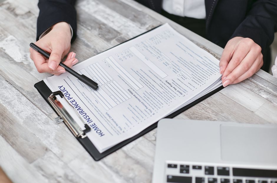 A person wearing a dark suit is seated at a wooden table, holding a black pen in their right hand and pointing it towards a home insurance policy document clipped to a black clipboard. Their left hand rests on the table, stabilizing the document, which contains printed text, checkboxes, and sections related to insurance details for house removals. The table has a weathered, light-colored wood surface with visible grain and slight distressing. To the right of the clipboard, part of an open silver laptop with a black keyboard is visible, positioned close to the edge of the image. The scene is lit with natural lighting, suggesting an indoor setting, possibly within a home or office environment associated with house relocation planning. As part of the moving process, the insurance document signifies the importance of securing appropriate liability coverage during furniture transport and packing in home relocations, which is a service offered by Man with Van Totteridge.