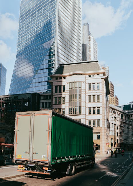A large green delivery truck with closed rear doors is parked on a city street next to tall, modern office buildings with glass facades. The truck is positioned in front of a historic multi-storey building with large windows and architectural detailing, indicating a busy urban environment suitable for home relocation and furniture transport. The scene shows the truck actively involved in a loading process, likely preparing for a house move or furniture delivery, with the surrounding area featuring pedestrians and vehicles, and natural daylight illuminating the buildings and street. Man with Van Totteridge, specializing in removals and moving services, appears to be coordinating this logistics operation within a city setting, utilizing appropriate equipment and vehicles for furniture transit and packing and moving activities.