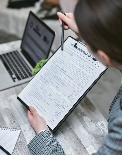 A person wearing a dark suit is seated at a wooden table, holding a black pen in their right hand and pointing it towards a home insurance policy document clipped to a black clipboard. Their left hand rests on the table, stabilizing the document, which contains printed text, checkboxes, and sections related to insurance details for house removals. The table has a weathered, light-colored wood surface with visible grain and slight distressing. To the right of the clipboard, part of an open silver laptop with a black keyboard is visible, positioned close to the edge of the image. The scene is lit with natural lighting, suggesting an indoor setting, possibly within a home or office environment associated with house relocation planning. As part of the moving process, the insurance document signifies the importance of securing appropriate liability coverage during furniture transport and packing in home relocations, which is a service offered by Man with Van Totteridge.